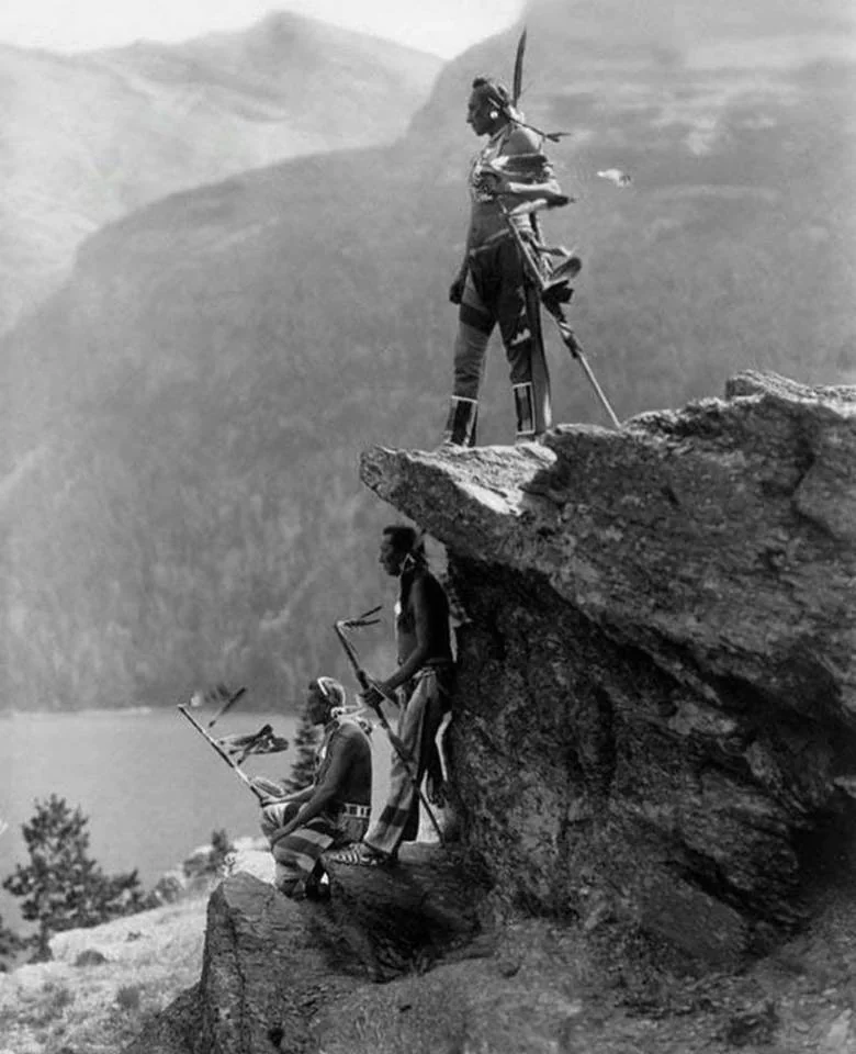 Three Native American men stand and sit on a rocky outcrop overlooking a distant lake and mountains. They hold traditional spears and wear native attire, including feathered headdresses. The scene is captured in a black and white photograph.