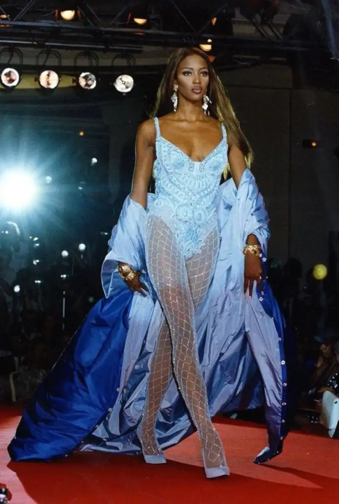 Model walking a runway in a light blue bodysuit with intricate detailing, paired with a long blue cape. She wears patterned sheer tights and high heels. The background features bright stage lights and a red carpet.