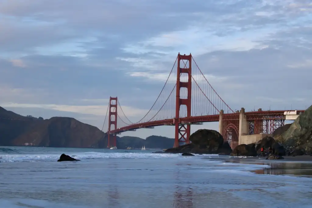 The image shows the Golden Gate Bridge spanning across San Francisco Bay. The iconic red suspension bridge is seen from a shoreline with calm waters and scattered rocks in the foreground, under a partly cloudy sky.