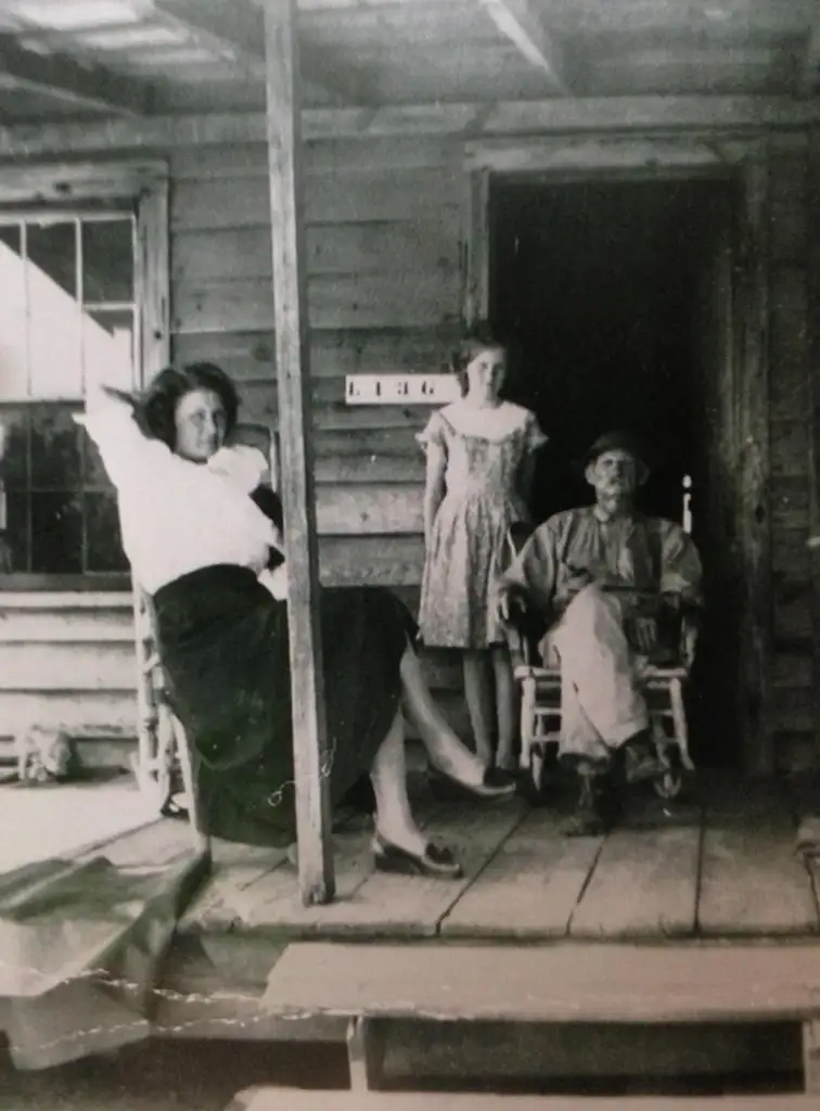 Three people on a wooden porch of a rustic cabin. One woman sits on the railing, another stands in the doorway, and an older man sits in a rocking chair. The cabin has wooden siding and a window with several panes.