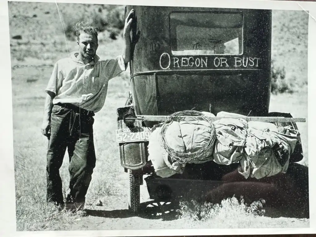 A man stands next to an old vehicle with a hand-painted "Oregon or Bust" sign on the back. The car is loaded with bags and supplies. The scene is outdoors in a rural setting, suggesting a journey or road trip.