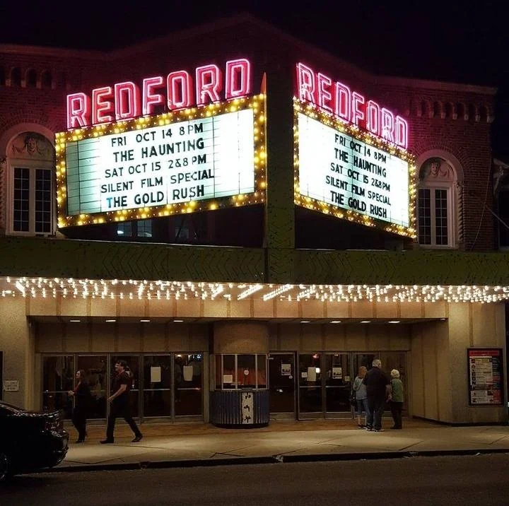 A vintage theater with a lit marquee reading "Redford" advertises upcoming screenings: "The Haunting" and "The Gold Rush" on October 14 and 15. People are walking by the entrance under string lights at night.