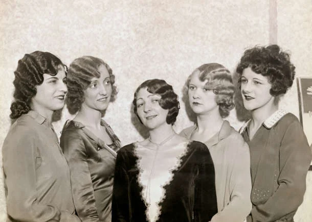 Five women in 1920s-style dresses and hairstyles stand together, smiling. The women are posed against a plain background. Their hairstyles are wavy and bobbed, and they wear a variety of satin and silk dresses.