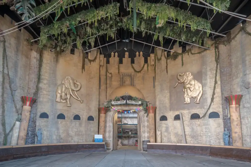 Abandoned amphitheater stage with elephants and castle-like decor. The ceiling is adorned with hanging plants. Bunting and a small cart are visible at the back. The scene has a dilapidated, overgrown appearance.