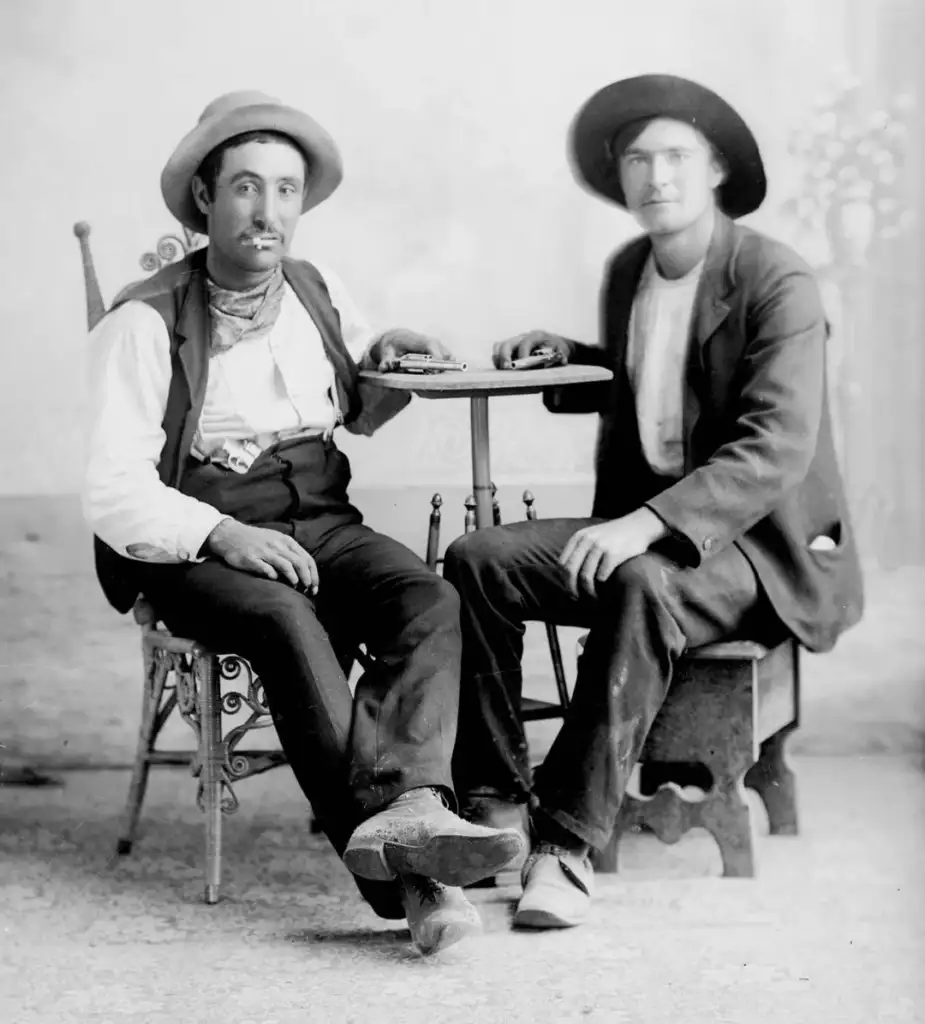 Two men sit in a vintage photograph. Both wear hats and suits; the man on the left has a mustache and crossed legs. They sit around a small table in a sparsely decorated room with a floral backdrop.