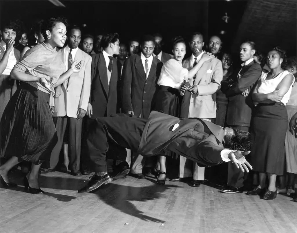 A dancer performs a dramatic backward lean in the middle of a circle of onlookers, all dressed in formal attire. The crowd appears captivated by the performance, with various expressions of amazement and excitement.