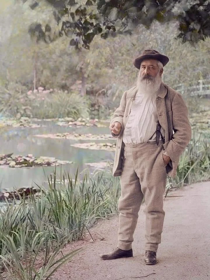 An elderly man with a long beard stands on a garden path near a pond filled with water lilies. He wears a suit, hat, and holds a pipe. Lush greenery and trees surround the tranquil setting.