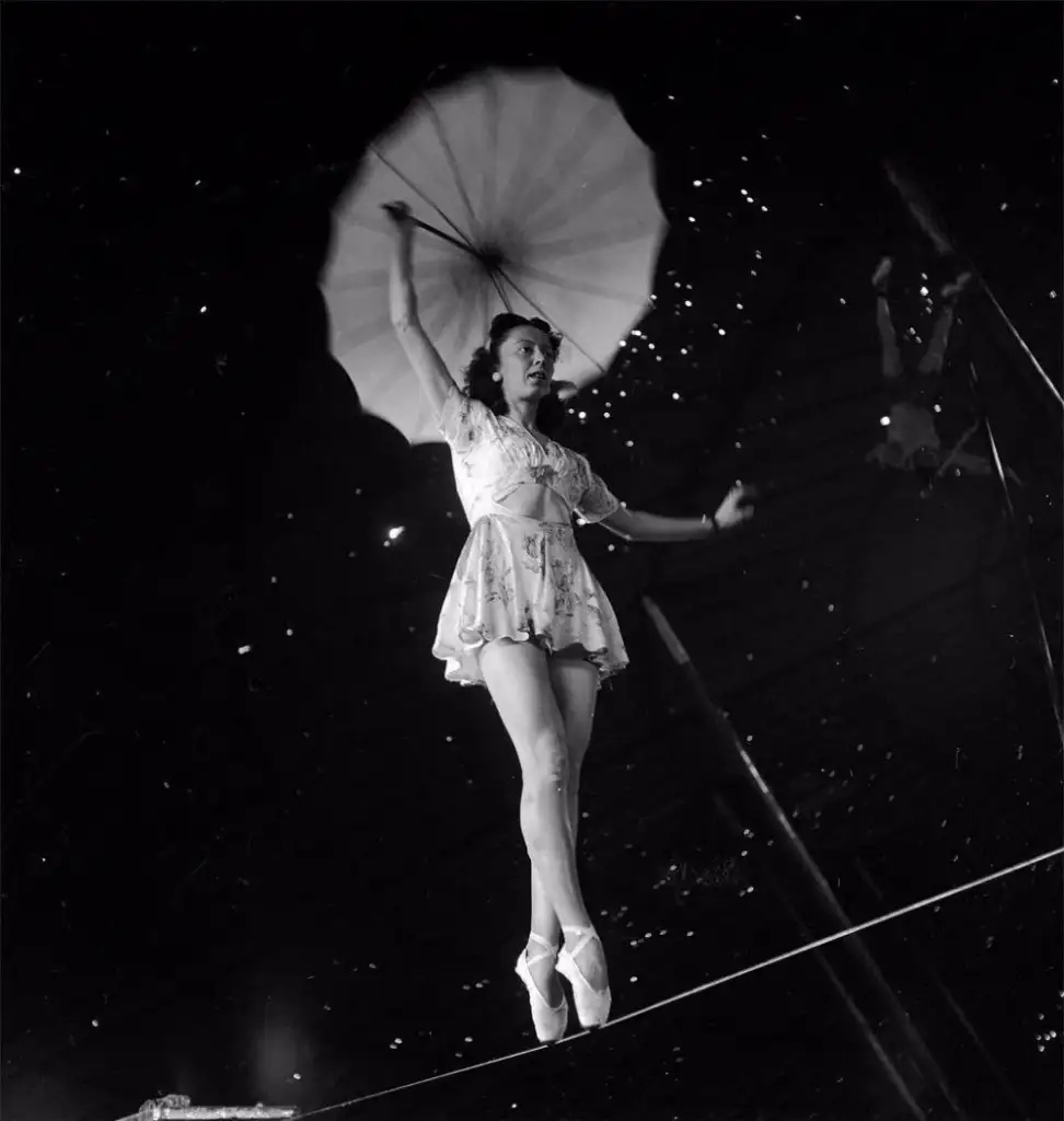 A performer in a short dress and ballet shoes gracefully balances on a tightrope, holding a large open parasol above her. The dark background highlights her poised stance and the umbrella.