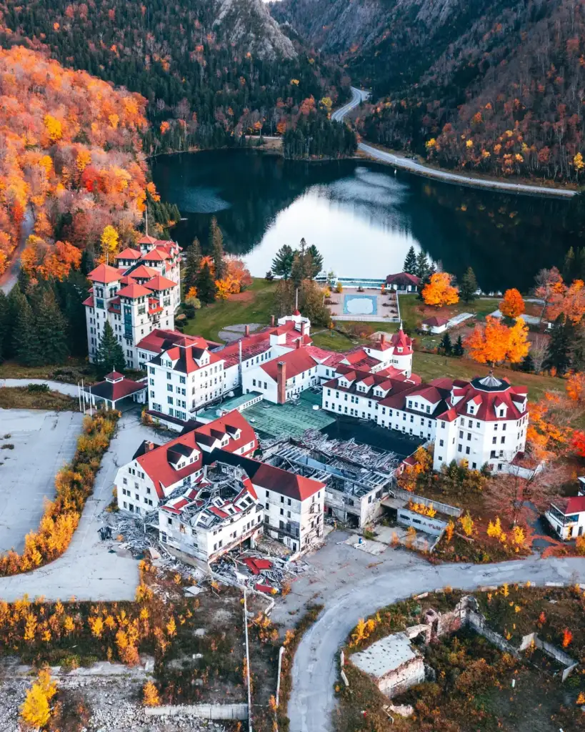 Aerial view of a cluster of large, historic buildings with red roofs set against an autumn backdrop. The structures are surrounded by colorful trees near a reflective lake. A winding road and mountains are visible in the background.