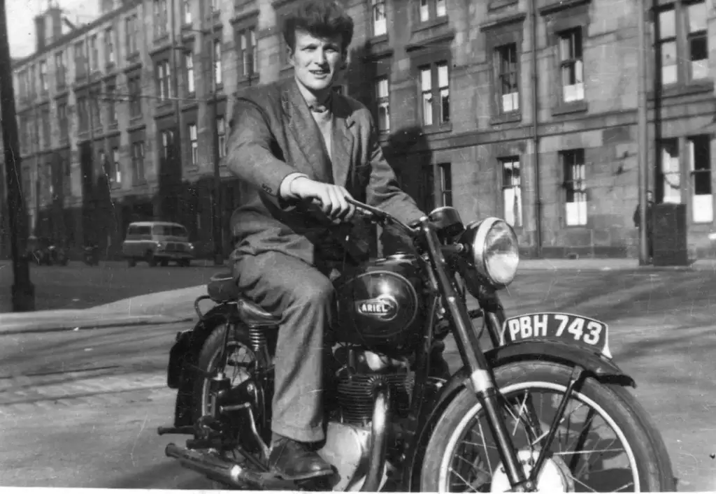 A person in a suit is riding a vintage Ariel motorcycle on a city street. The background shows old buildings and a parked car. The motorcycle has a visible license plate with the code "PBH 743." The photo appears to be black and white.