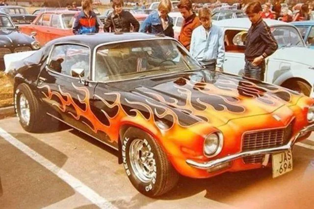 A vintage car show with people gathered around a classic car. The vehicle is painted with a flame design, blending orange, red, and black, and has a shiny finish. Other cars and attendees are visible in the background.