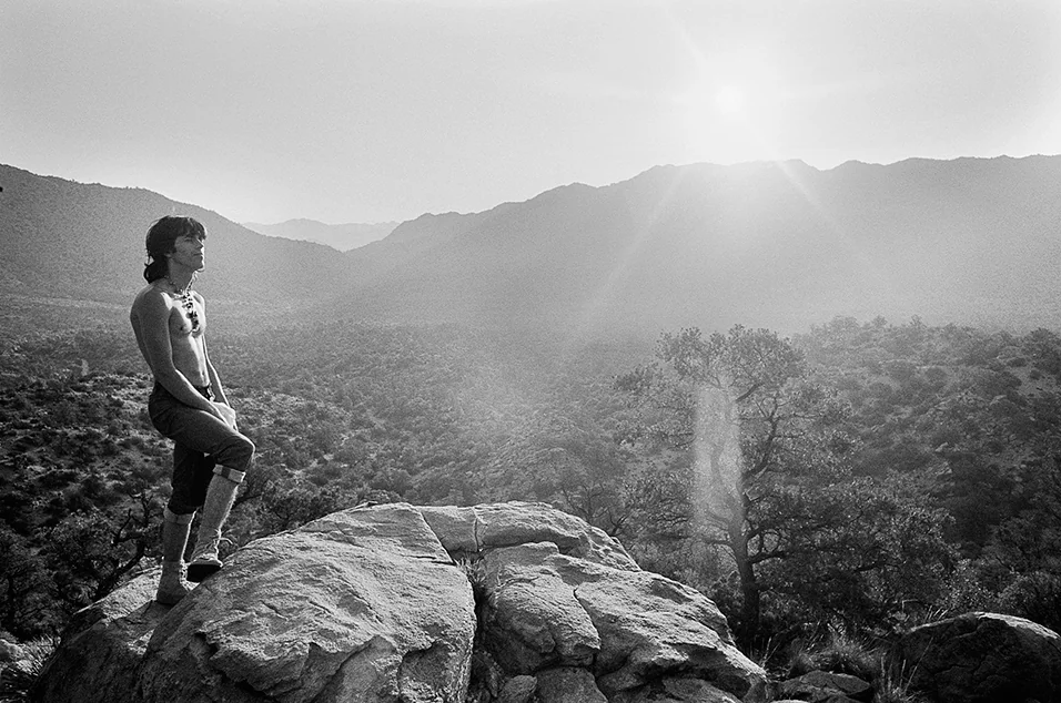 A person standing on a large rock looks out over a sunlit valley with scattered trees and distant mountains. The sun is low in the sky, casting a soft glow and creating a serene atmosphere. The photo is in black and white.