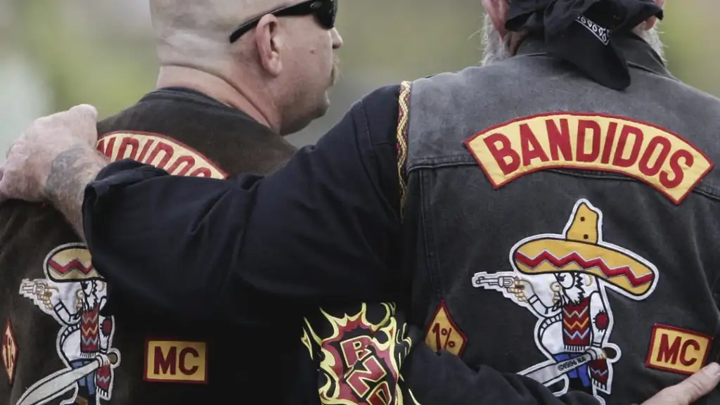 Two people with arms around each other wear "Bandidos MC" motorcycle club jackets. The jackets feature a cartoon character wearing a sombrero and holding a gun. Both have their backs to the camera.