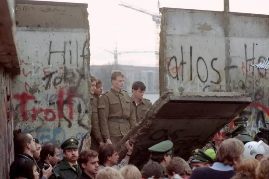 A crowd gathers as a large section of graffiti-covered concrete is removed from a wall. Several people stand on the debris, while others watch. Construction cranes are visible in the background. The scene is bustling with activity and excitement.