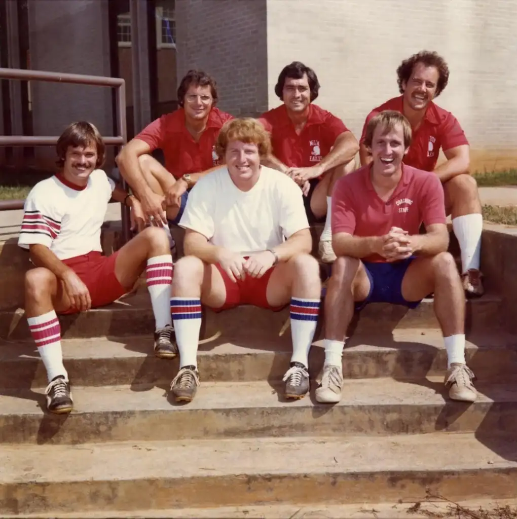 Six men in athletic clothing sit on outdoor steps. Three in white shirts and three in red, with matching shorts and striped socks. They smile at the camera. A building and handrail are visible in the background.