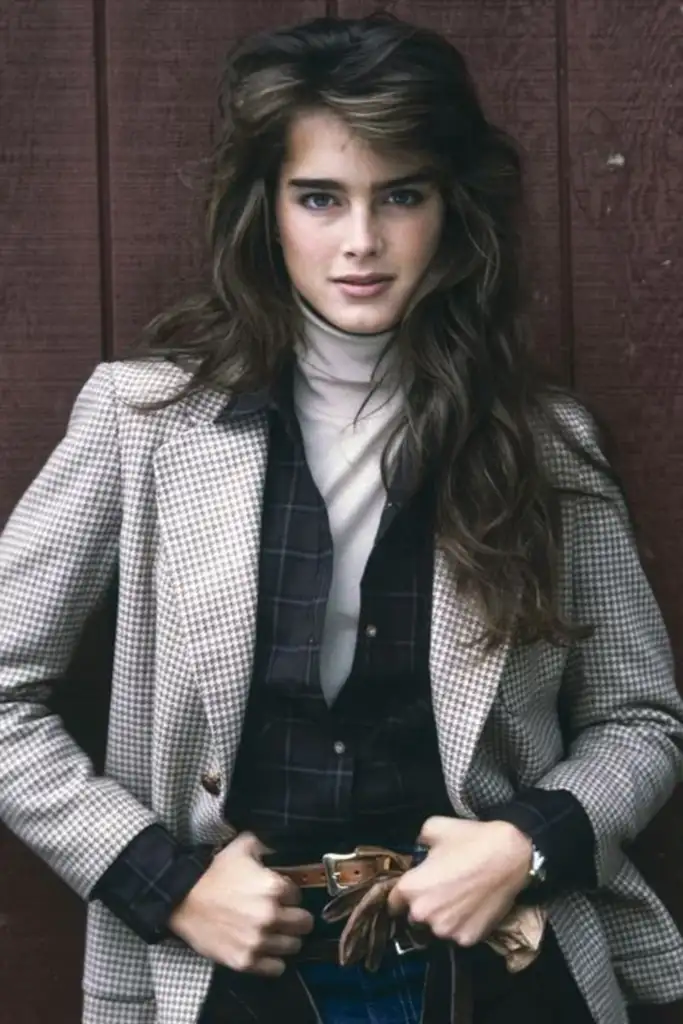 A person with long, wavy brown hair stands against a wooden wall. They wear a checkered blazer over a black shirt and a light turtleneck. Their hands are in the pockets of blue jeans, and they have a confident expression.