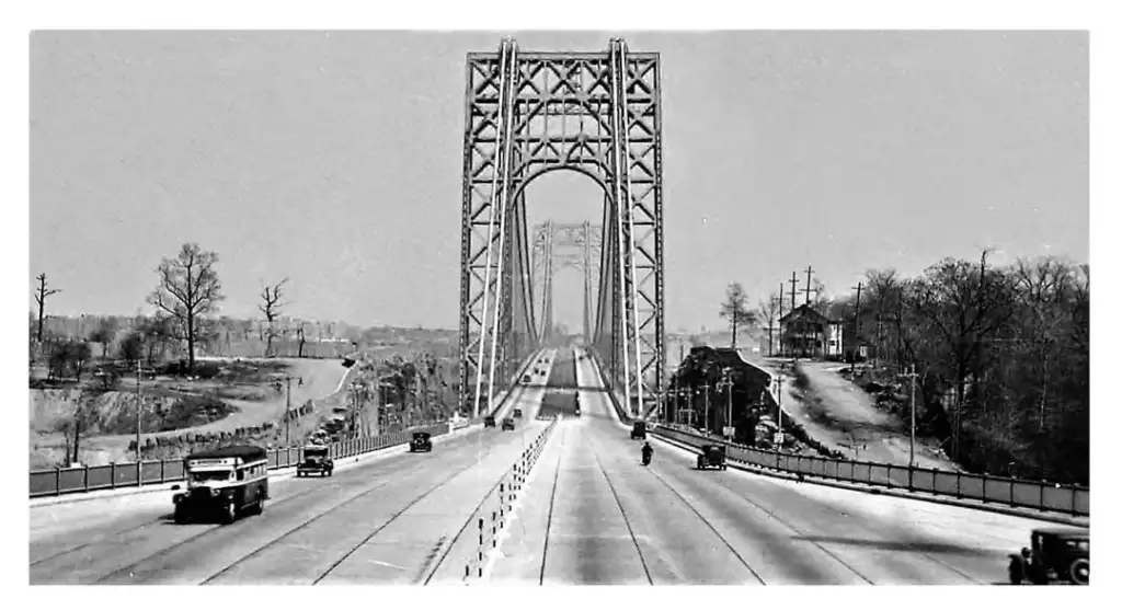 A black and white photo of an old suspension bridge spanning a river. Cars and a motorcycle are driving across, with leafless trees and utility poles lining the road. The bridge's massive metal structure towers overhead.