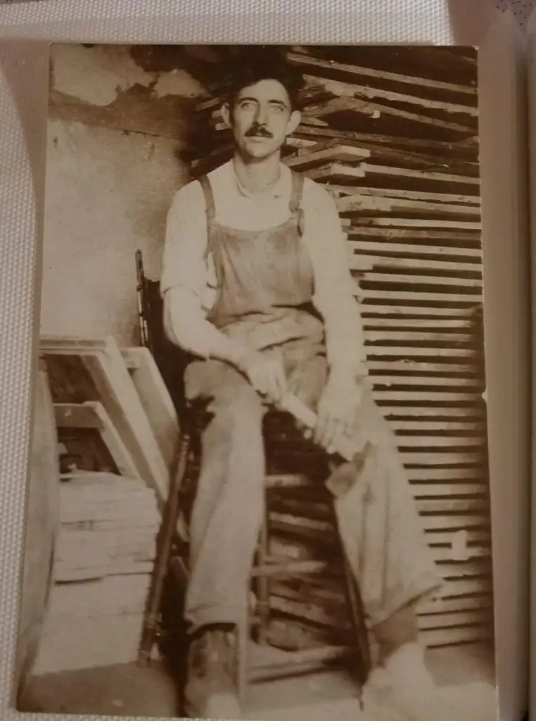 A sepia-toned photo of a man with a mustache sitting on a chair. He is wearing overalls and a long-sleeve shirt. Behind him are neatly stacked wooden frames or planks. The setting appears to be a workshop or storage area.