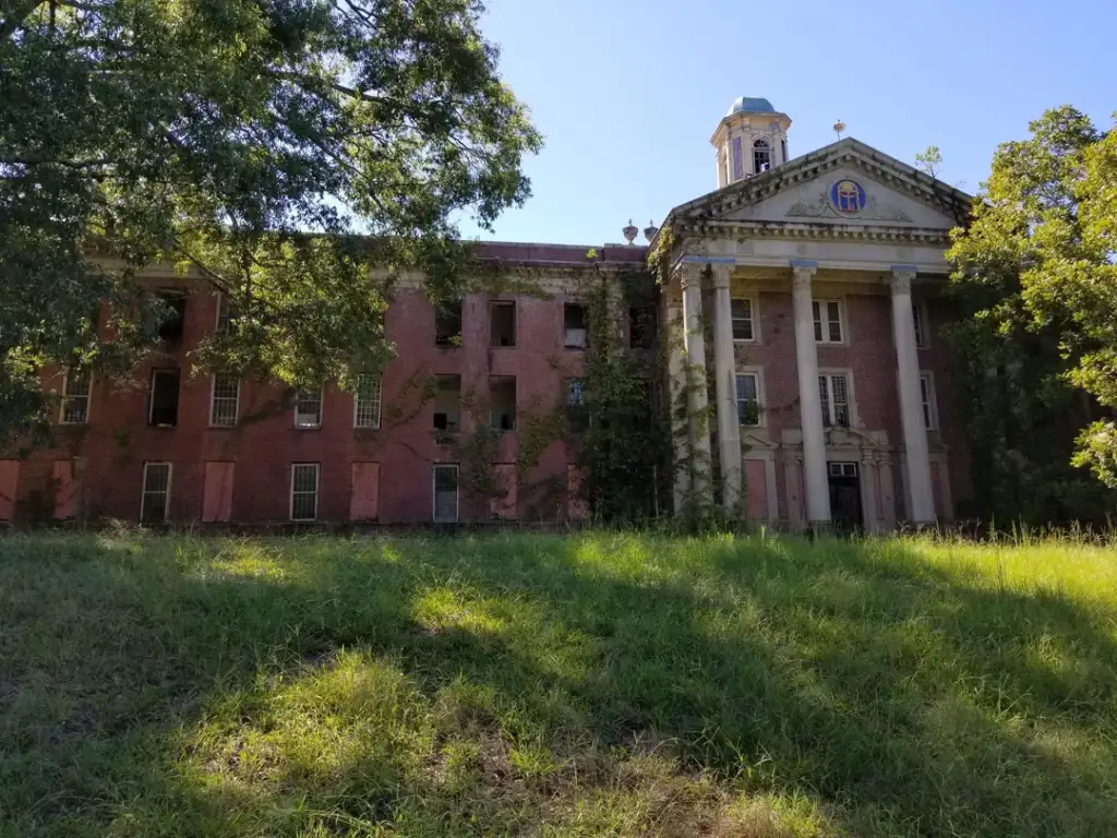 An abandoned, red-brick building with a classical columned entrance and a small dome on top. The structure is partially covered with overgrown vines, surrounded by green grass and large trees under a clear blue sky.