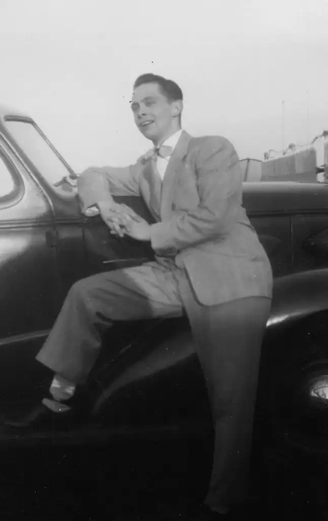 A man in a suit leans on the front of a vintage car, smiling. He is holding a pipe and wearing glasses, with one foot resting on the car's fender. The setting is outdoors during the day.