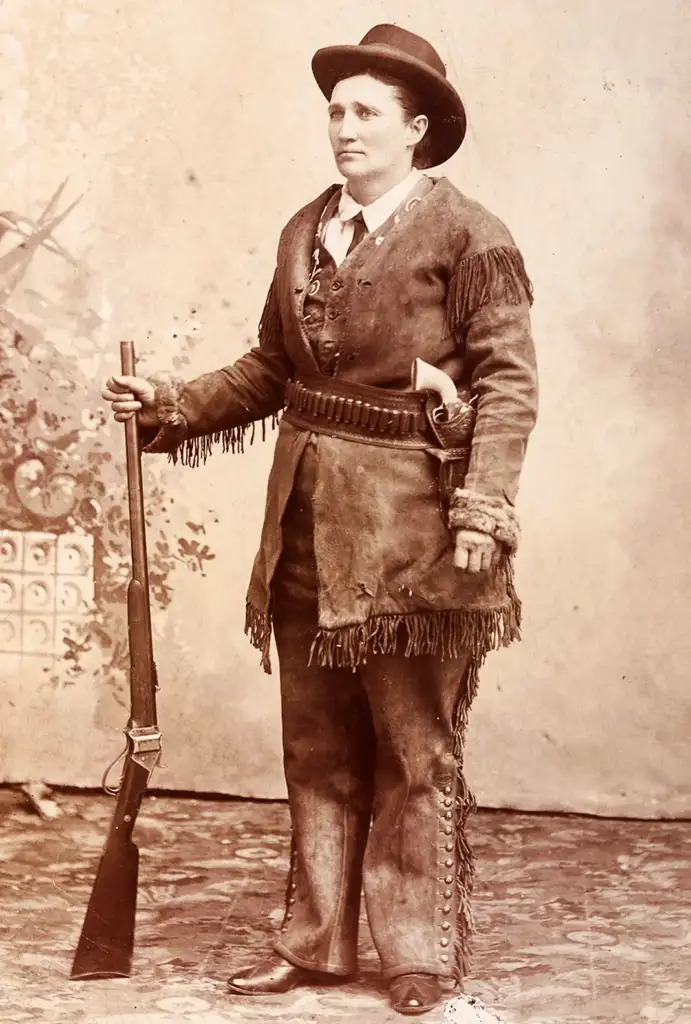 A person in vintage Western attire stands holding a rifle. They wear a wide-brimmed hat, fringed jacket, and pants with buttons down the leg, along with a belt featuring bullets and a holstered gun. The backdrop is a decorative, rustic scene.