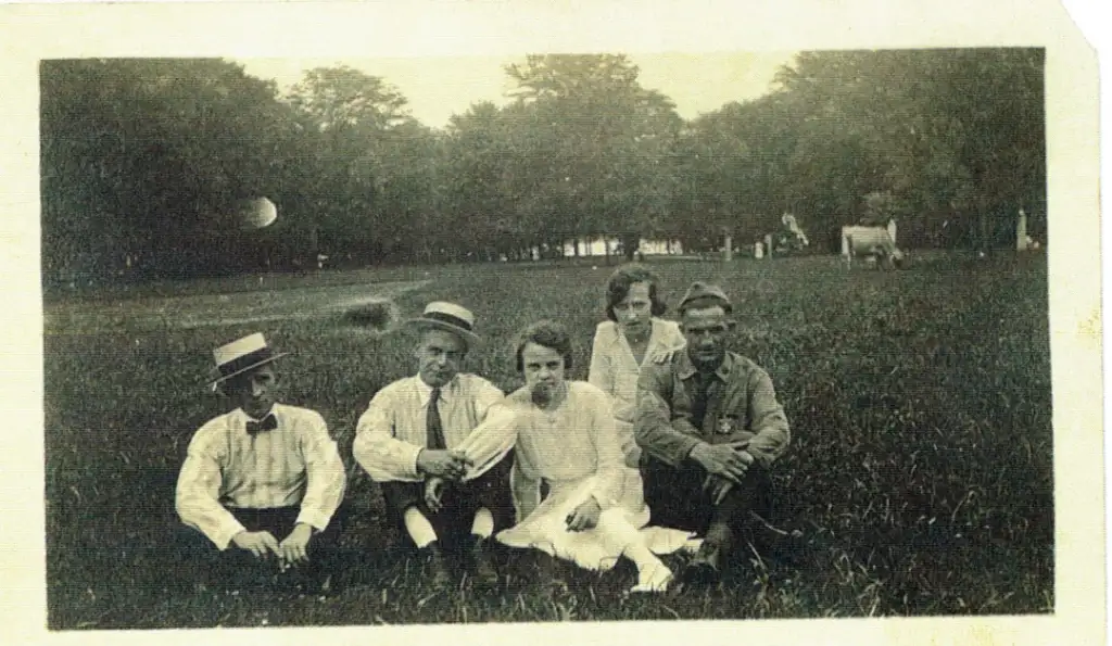 A vintage photo of five people sitting on grass in a park. Four men and one woman are dressed in early 20th-century clothing. Trees are in the background, with a cow grazing on the right. A faded moon appears in the sky.