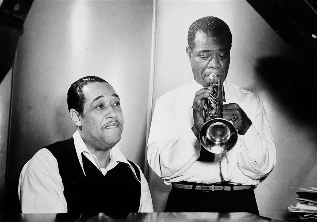 A black and white image of two musicians: one seated and smiling, wearing a vest over a shirt, while the other stands playing a trumpet, dressed in a white shirt. They are both focused on the music, set against a plain background.