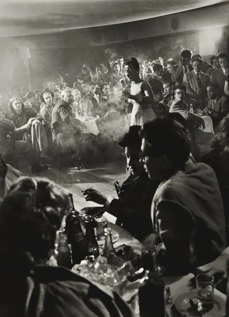 A woman in a white dress sings on stage at a smoky, crowded nightclub. People sit at tables with drinks, watching her perform. The audience is a mix of men and women, all focused on the performance in the dimly lit room.