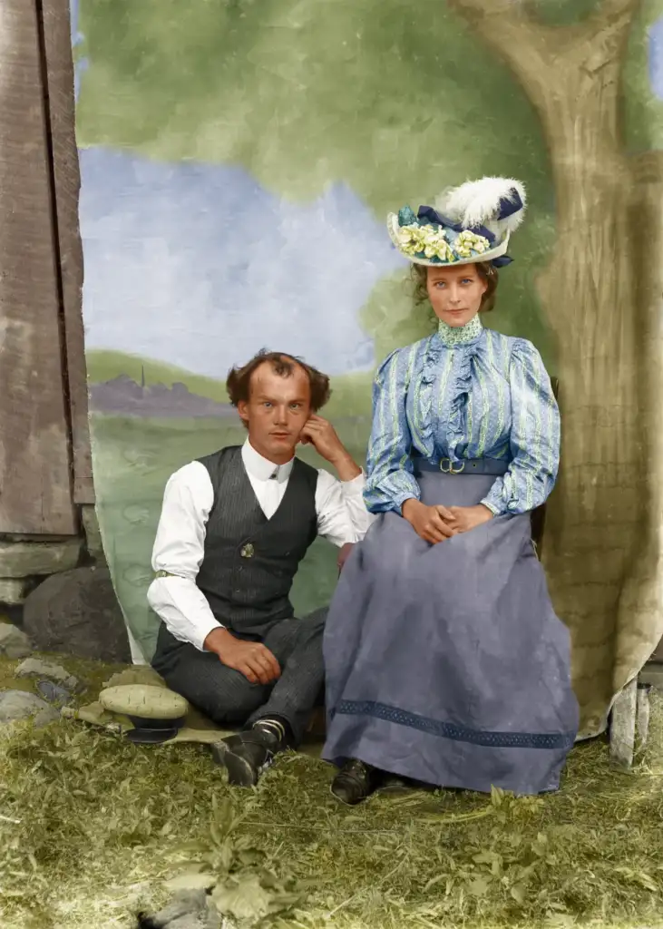 A man and woman in early 20th-century attire sit outdoors against a painted backdrop. The man wears a vest and tie, and the woman dons a hat with feathers and a long skirt. They pose on grass beside a painted tree and landscape.