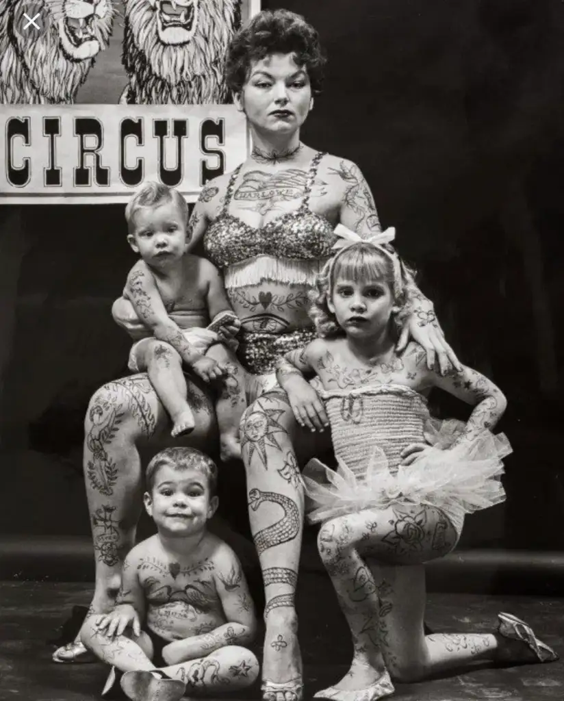 A tattooed woman poses with three children, all with body art. They are in front of a backdrop with the word "Circus" and lion illustrations. The children wear various outfits, adding a playful yet bold contrast to the scene.