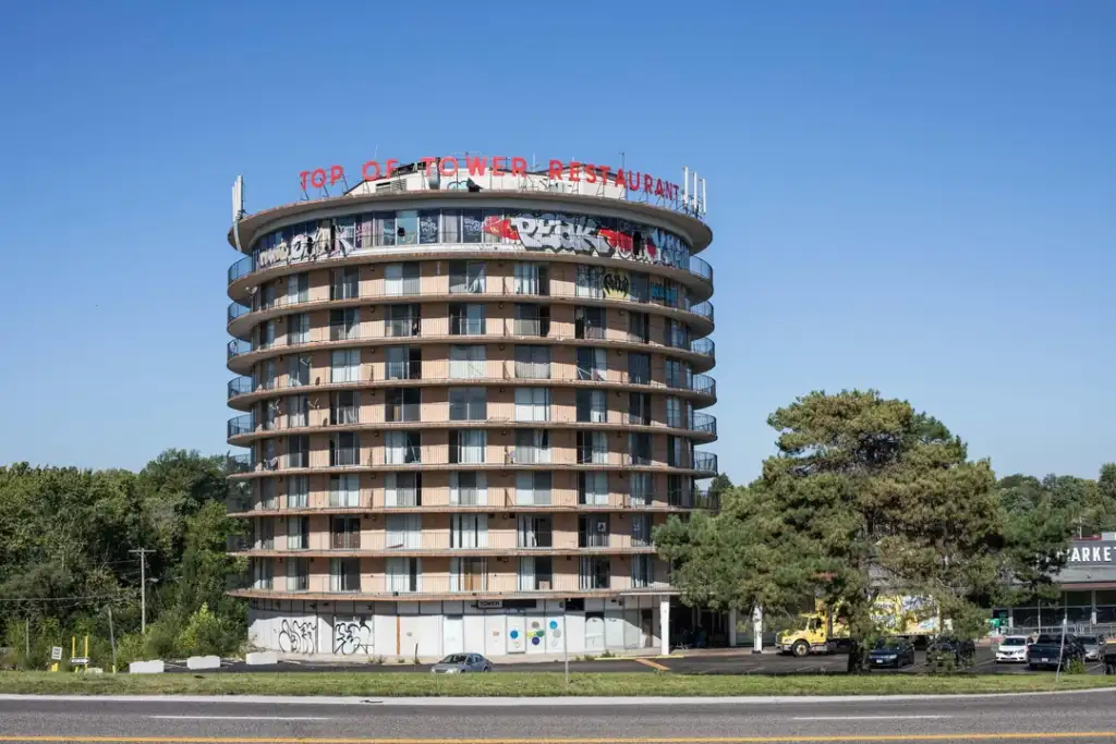 Round, multi-story building with balconies, featuring graffiti on top. A red sign reads "Top of Tower Restaurant." Trees and a road with a yellow line are in the foreground, under a clear blue sky.