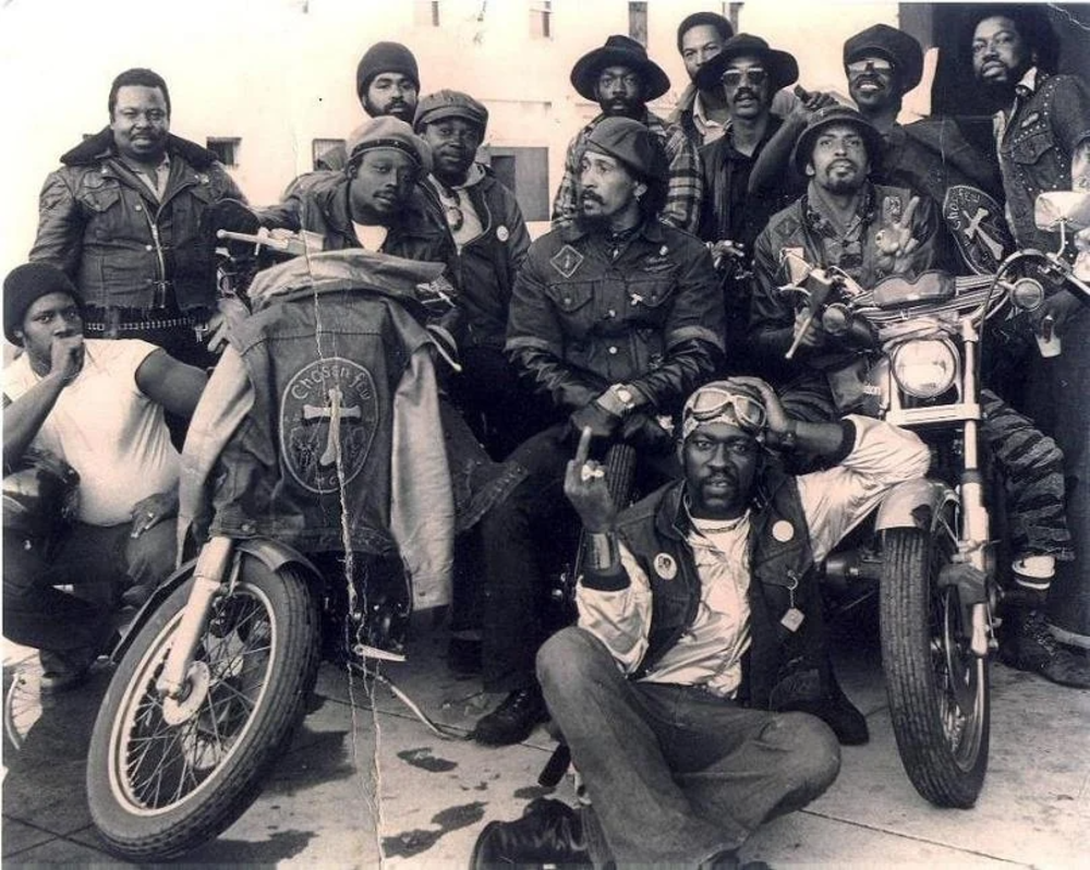 A group of eleven men wearing leather jackets and hats, posing together with two motorcycles. The men display various expressions and poses, with some wearing patches or badges. The scene is in black and white, suggesting a vintage photo.