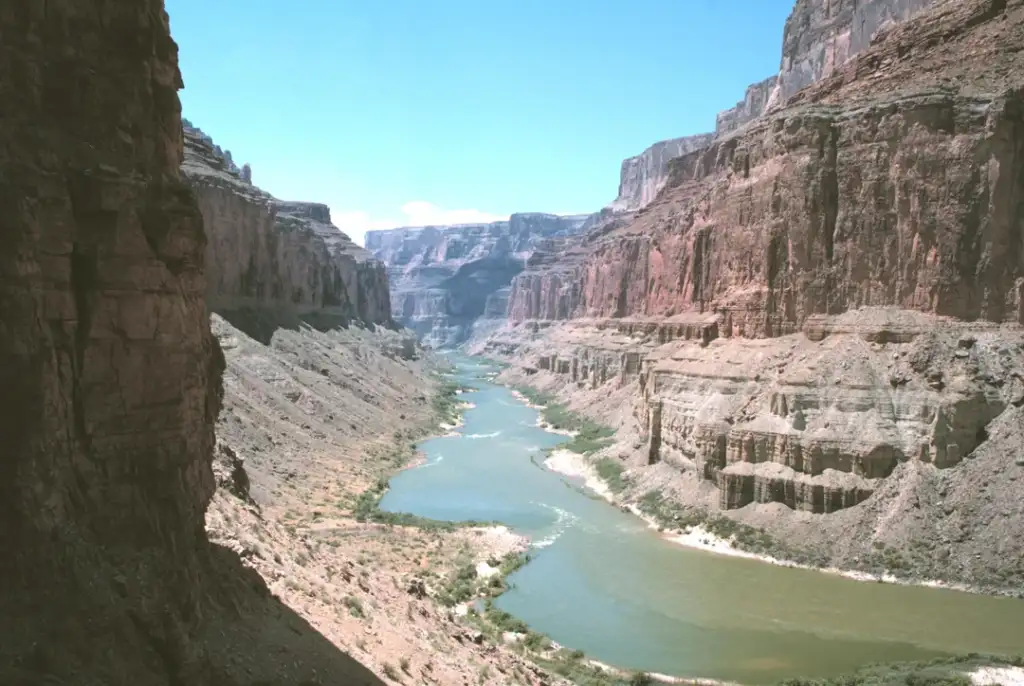 A vast canyon landscape with towering red rock cliffs on both sides and a river winding through the center. The sky is clear and blue, highlighting the rugged textures of the canyon walls.