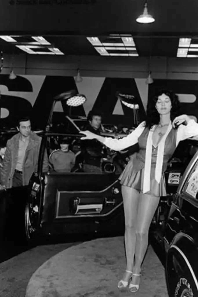 A woman poses in front of a Saab car with an open door at a car show. She is smiling, wearing a short outfit and holding a microphone. Several people are in the background, and the Saab logo is visible on the wall. The image is in black and white.