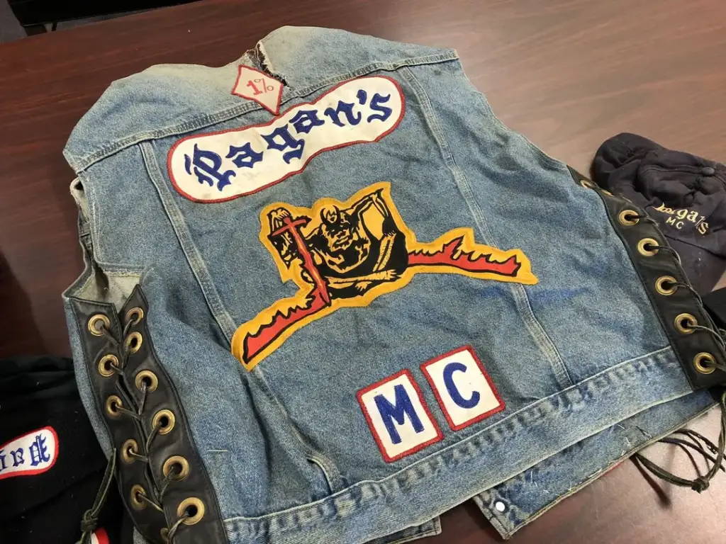 Denim biker vest with "Pagan's MC" patches on a table. The center design features a figure holding a staff on a dragon, with a "1%" symbol above. A black cap and glove with similar symbols are nearby.