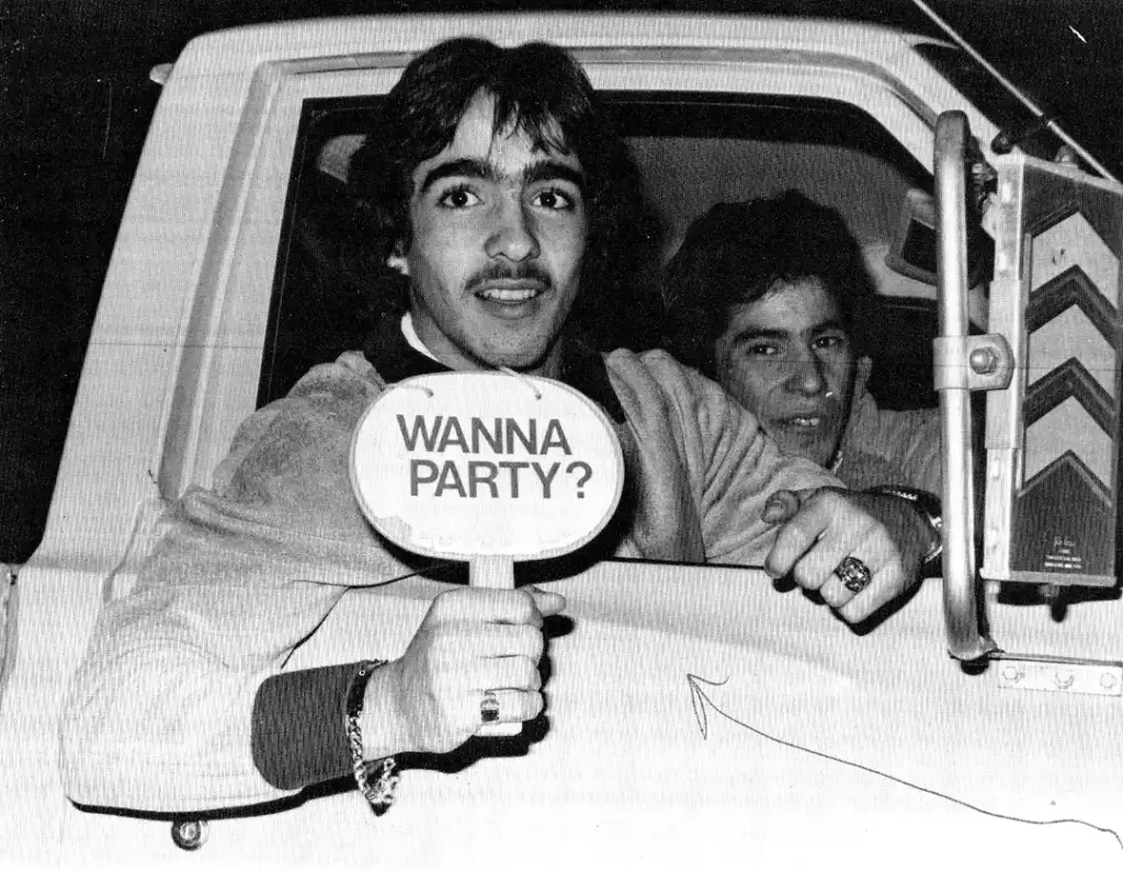 Two people in a vehicle, with one holding a circular sign reading "WANNA PARTY?" They appear excited and are looking out the window. The image is in black and white.