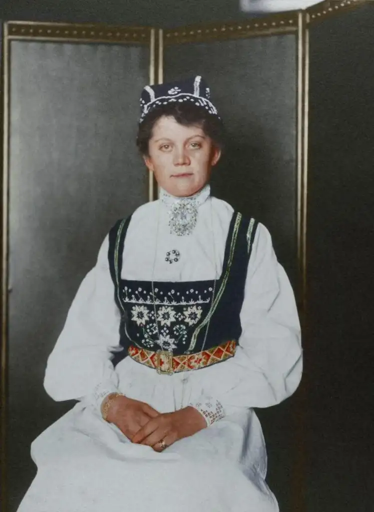 A woman sits wearing a traditional Scandinavian folk costume with detailed embroidery. The outfit includes a white blouse, dark vest, and a matching cap. She poses with hands neatly folded, against a plain, dark background.