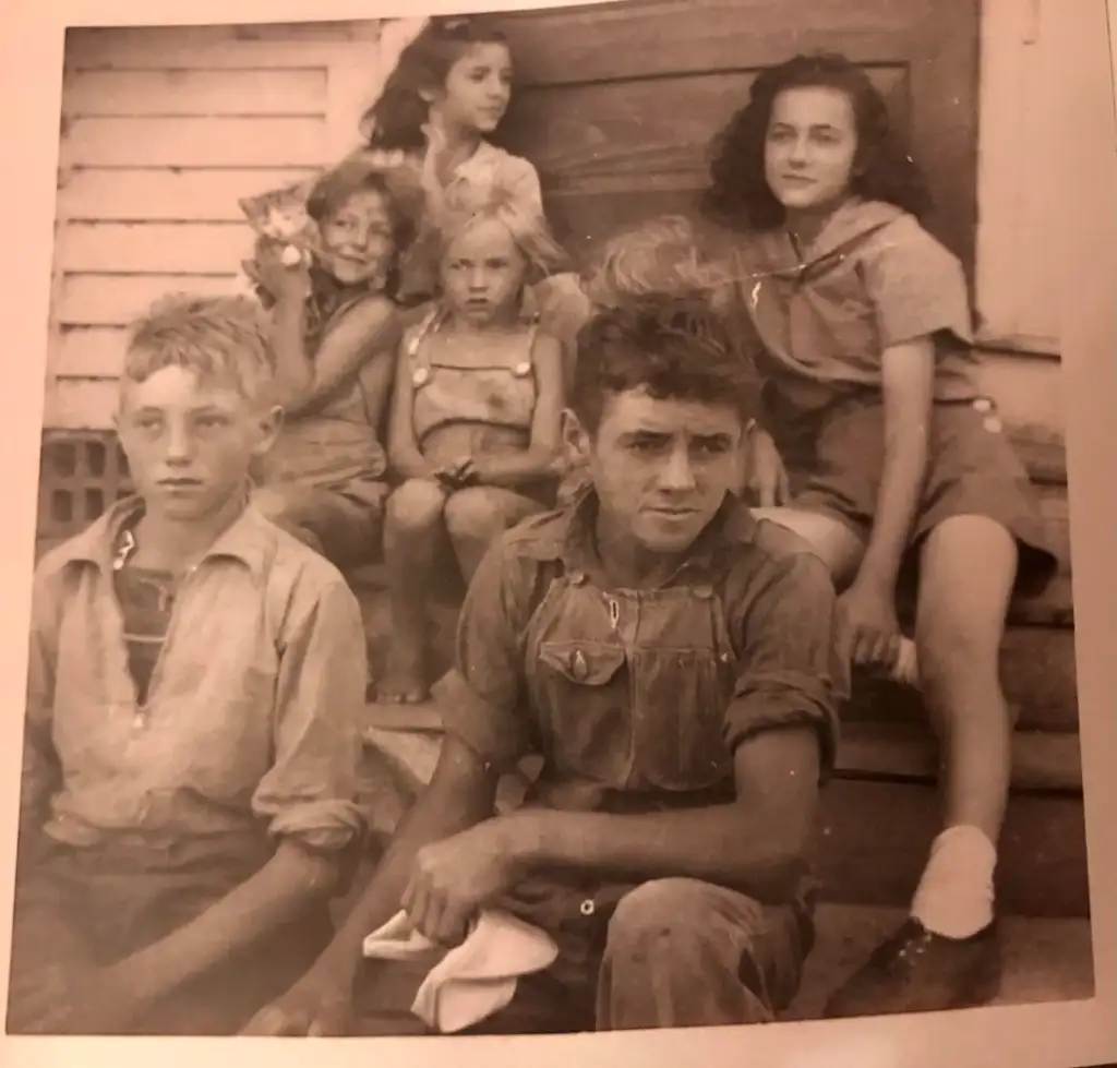 A black and white photo of six children sitting on wooden steps. Three girls and three boys, all in casual clothes from an earlier era. Some have short hair, while others have wavy hair. The expressions vary, with a mix of smiles and serious looks.