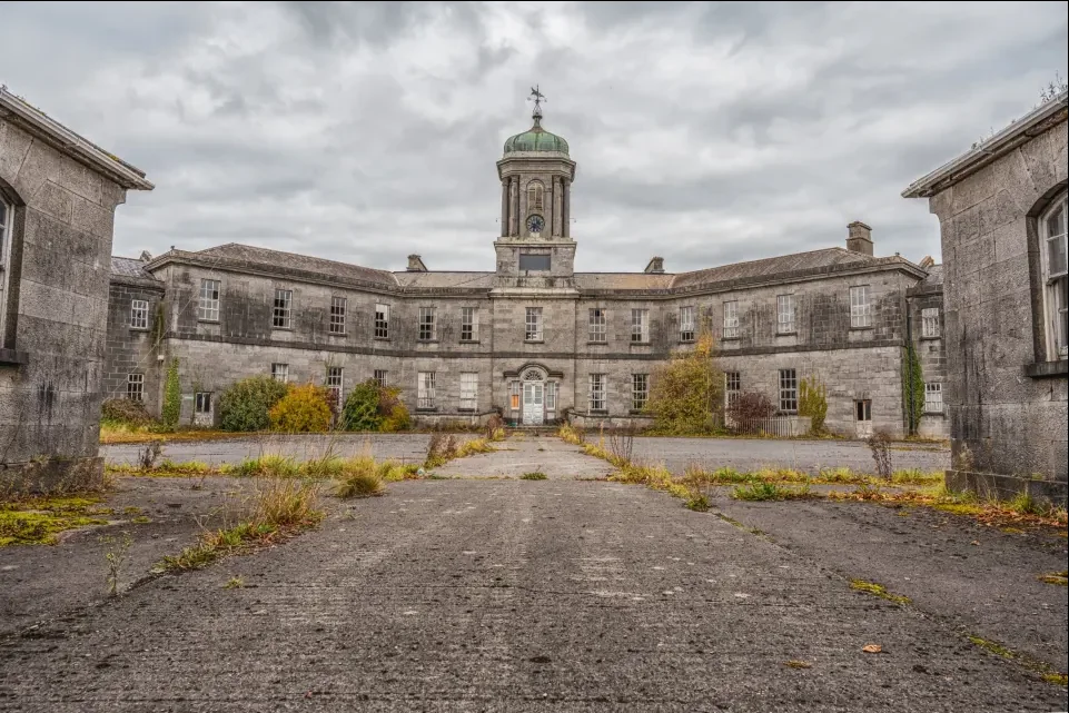 A large, abandoned stone building with a central tower and dome sits under an overcast sky. The courtyard is overgrown with weeds and vegetation. The structure shows signs of aging and neglect, with a symmetrical design and multiple window panes.