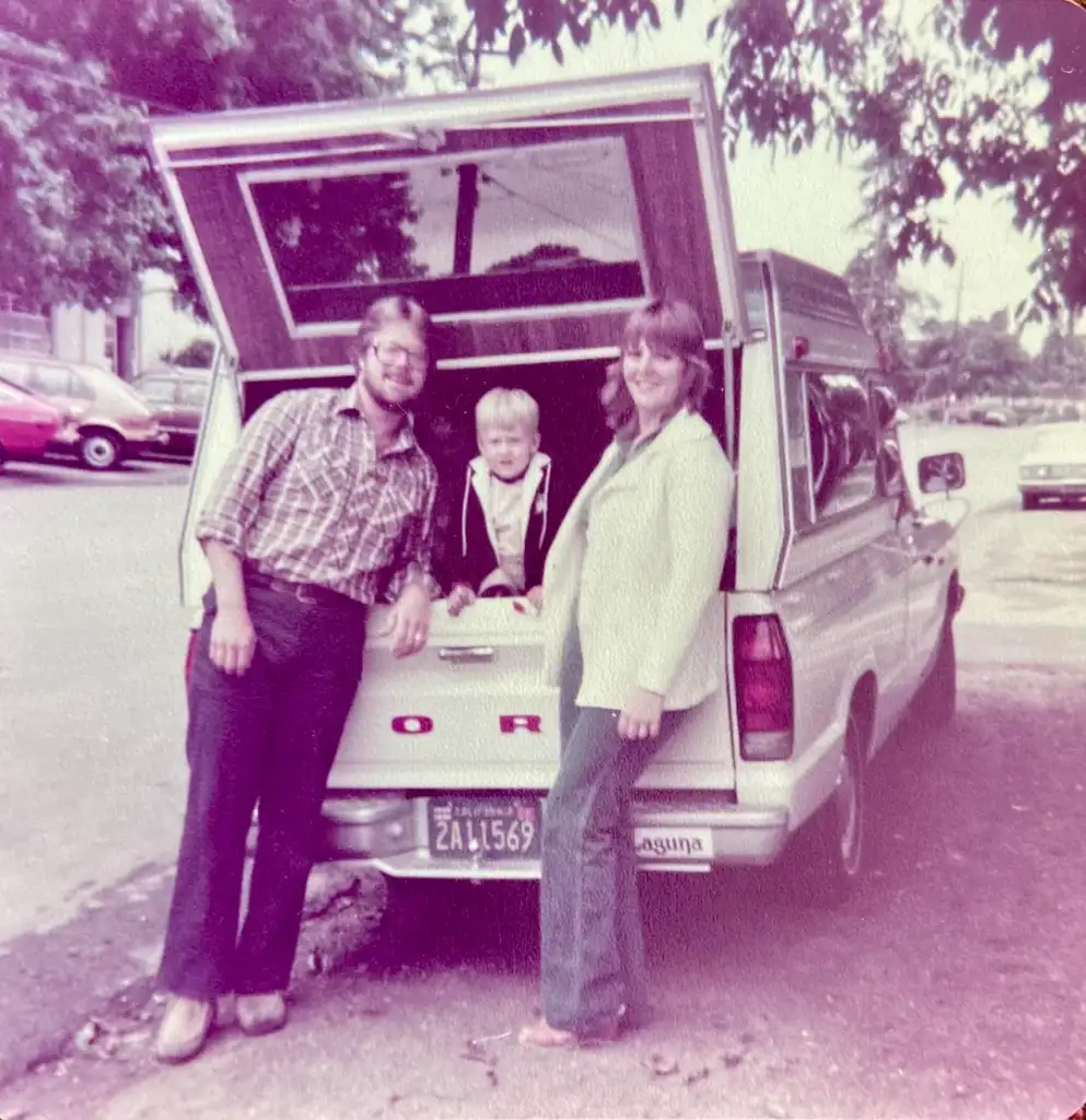 A man, child, and woman smile at the camera while leaning against the open tailgate of a white Ford pickup truck. Trees and parked cars line the street in the background. The image has a vintage look.