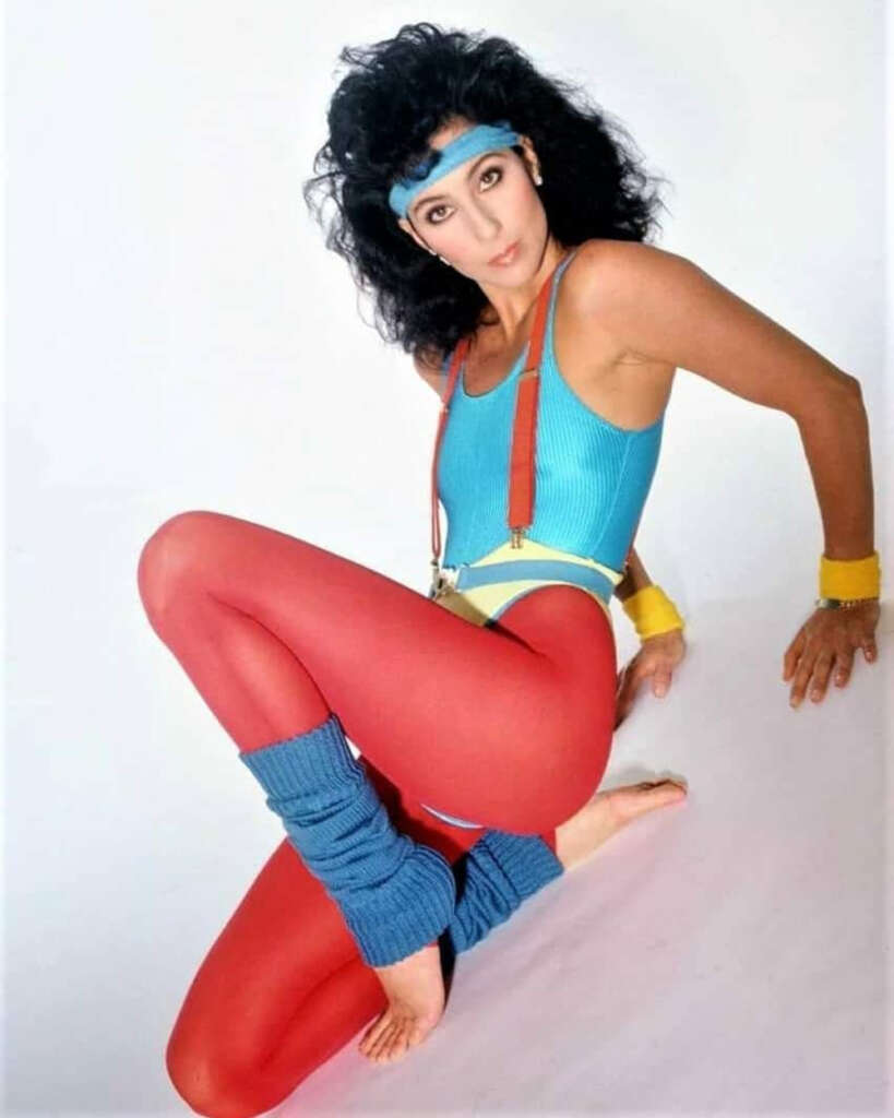 Person in 1980s workout attire, featuring a blue leotard, red leggings, blue leg warmers, and yellow wristbands. They wear a blue headband and have curly dark hair, posing against a white background.