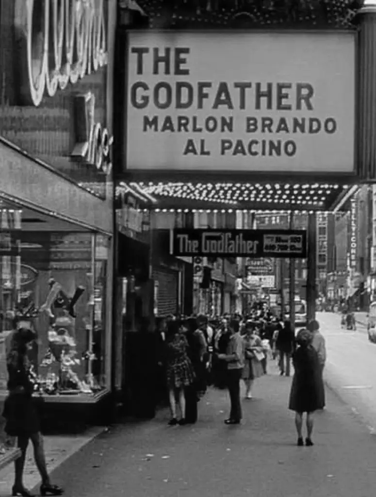 Black and white photo of a cinema marquee displaying "The Godfather" with Marlon Brando and Al Pacino. People are walking and standing near the entrance, and storefronts are visible along the street.