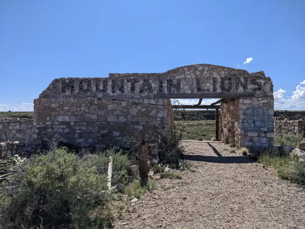 Stone ruins of a building stand under a clear blue sky. The structure has a partial sign reading "MOUNTAIN LIONS" and is surrounded by sparse vegetation and a gravel path leading through the archway.