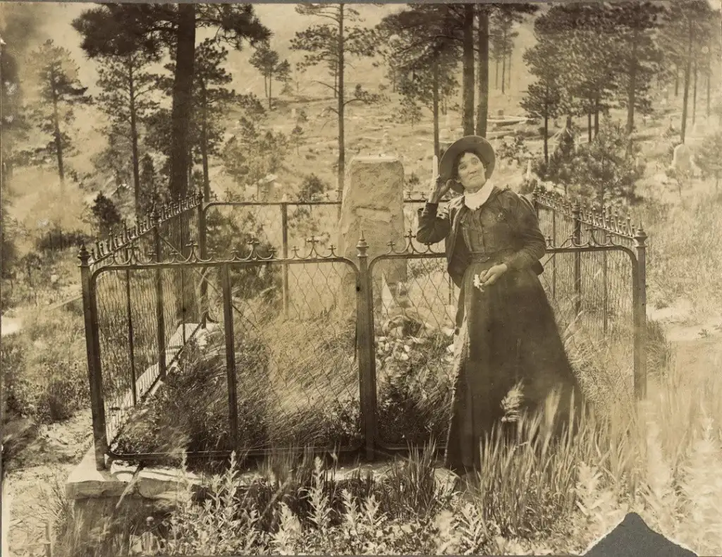 A woman in early 20th-century attire stands beside a fenced grave in a forested cemetery. She wears a long dress, hat, and holds an object. The background features trees and a slightly hilly landscape, creating a serene, historical scene.