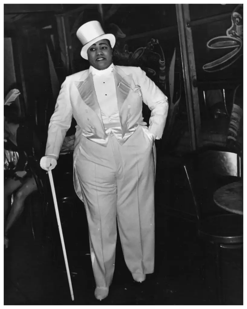 Person in a white tuxedo and top hat, holding a white cane, standing in a dimly lit room with chairs and abstract wall art.