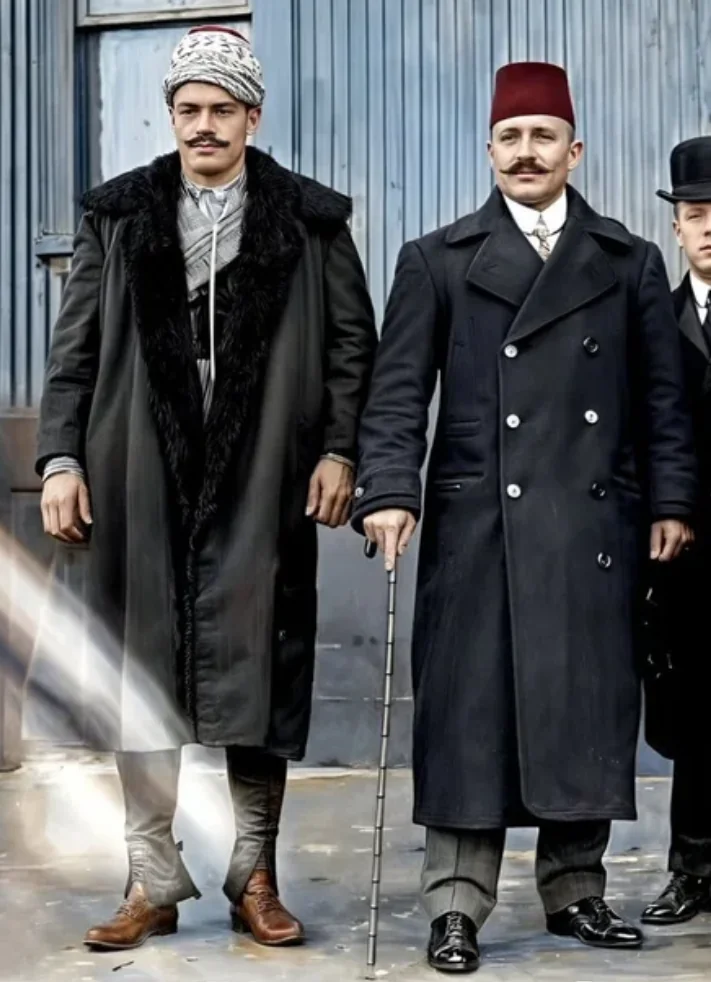 Two men in early 20th-century attire stand side by side. The man on the left wears a fur-lined coat and patterned headwear, while the man on the right wears a double-breasted coat, fez, and holds a cane. A third person is partially visible.