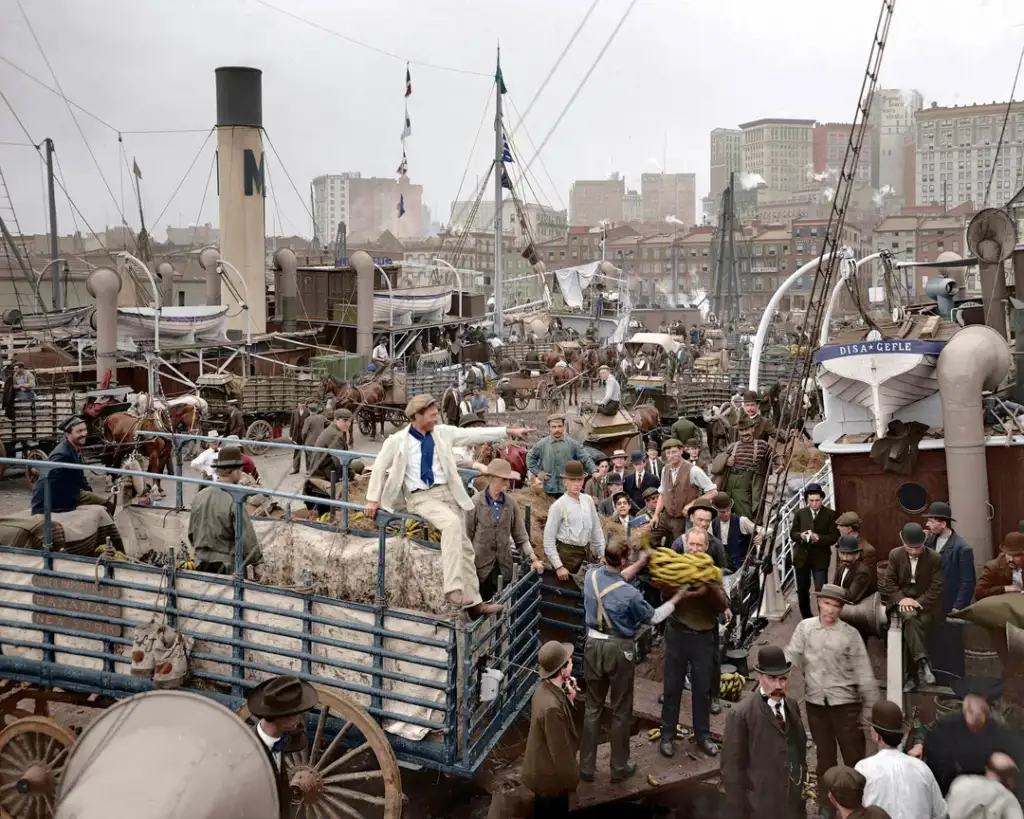 A bustling dock scene with workers loading goods onto horse-drawn carts and ships. Men in hats and suits oversee the activity. Smoke rises from ship funnels, with city buildings and a cloudy sky in the background.