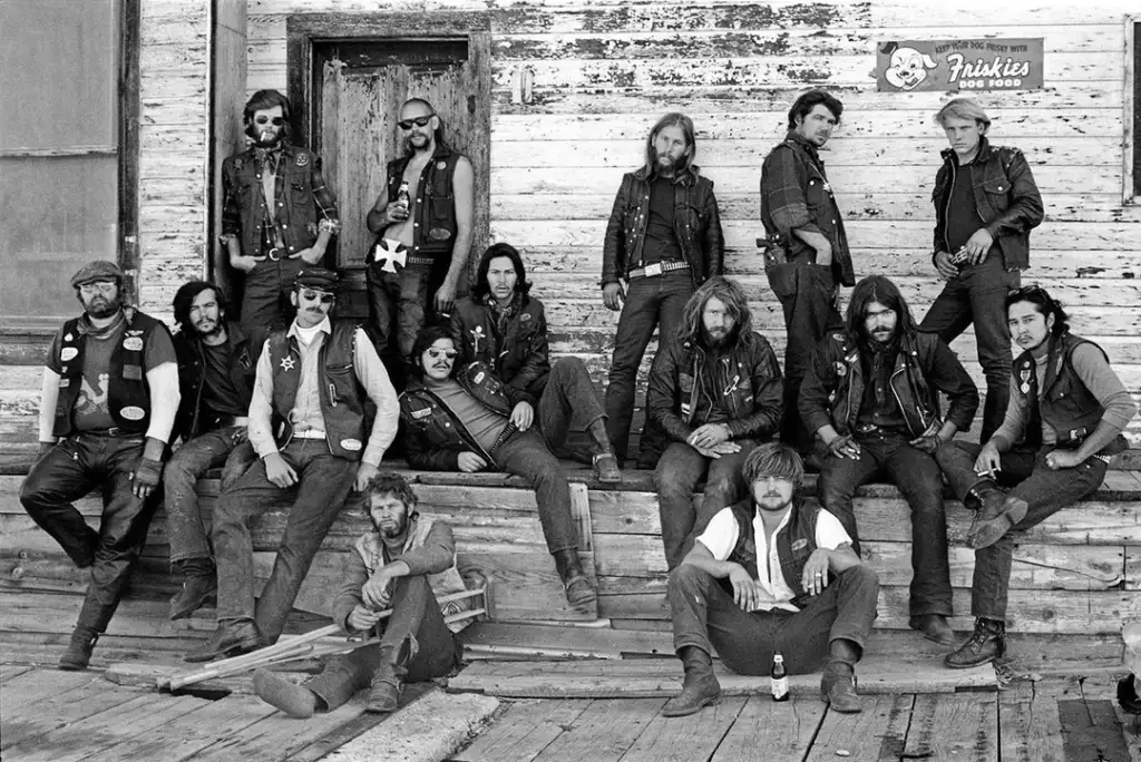 Black and white photo of a group of leather-clad bikers posing in front of a worn wooden building. Some sit on the porch steps, while others stand or lean against the walls. A "Friskies" sign is visible above the door.