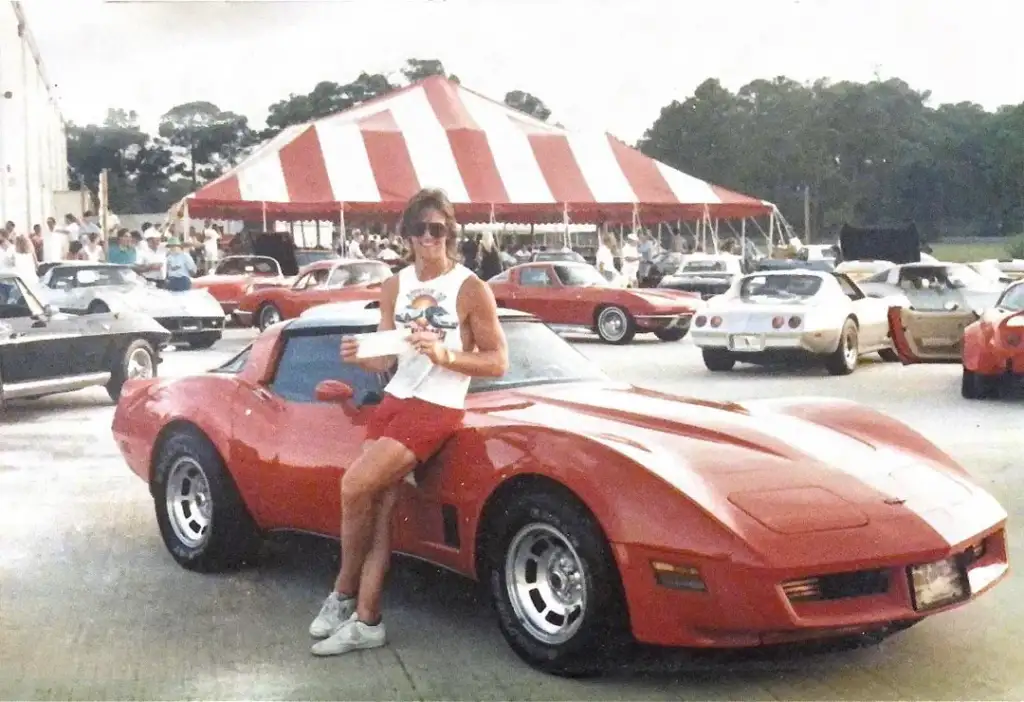 A person in a tank top and shorts leans against a red sports car, holding a drink. Behind them is a large red and white striped tent and a crowd of people. Several other cars are parked nearby. The setting appears to be an outdoor event.