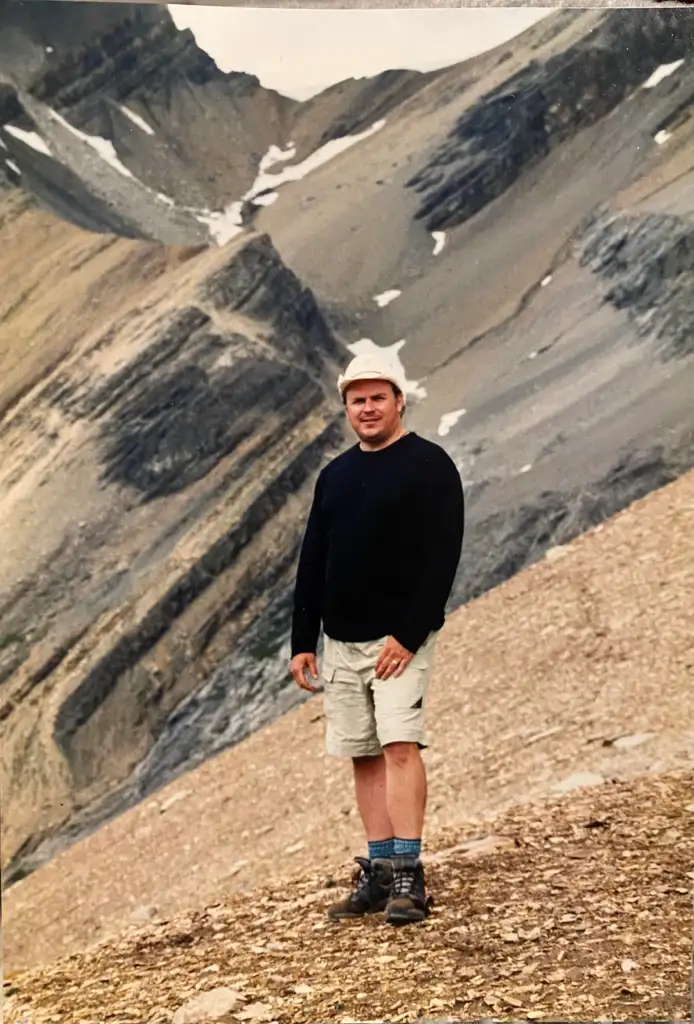 A person wearing a white hat, black sweater, light-colored shorts, and hiking boots stands on a rocky mountain slope. The background features rugged mountain terrain with patches of snow and cloudy skies.